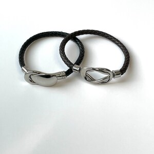 May include: Two braided leather bracelets. One is dark brown, the other is black. Each bracelet has a silver-colored clasp. One clasp is a solid oval, the other is a twisted knot design.