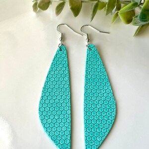 May include: A pair of turquoise earrings with a honeycomb pattern. The earrings are teardrop-shaped and hang from silver-colored hooks. The earrings are displayed on a white surface with green foliage in the background.