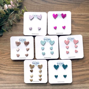 May include: Set of six pairs of heart-shaped earrings in various colors, including pink, teal, gold, and light blue. Each pair features three different sizes of hearts.