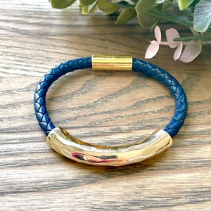 May include: A navy blue leather bracelet with a gold magnetic clasp. The bracelet is braided and has a curved gold bar in the centre.