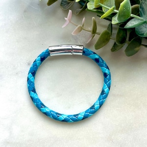 May include: Braided blue leather bracelet with a silver magnetic clasp. The bracelet features a mix of light and dark blue tones. The accessory is displayed on a white marble surface with green foliage in the background.