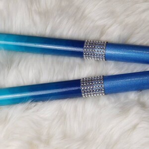 May include: Two cylindrical drumsticks featuring a smooth gradient finish that transitions from light sky blue to a deep royal blue. Each stick is accented with a central band of sparkling silver rhinestones that wrap around the middle section.