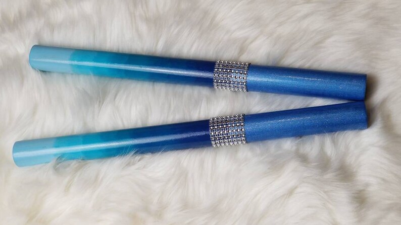 May include: Two cylindrical drumsticks featuring a smooth gradient finish that transitions from light sky blue to a deep royal blue. Each stick is accented with a central band of sparkling silver rhinestones that wrap around the middle section.