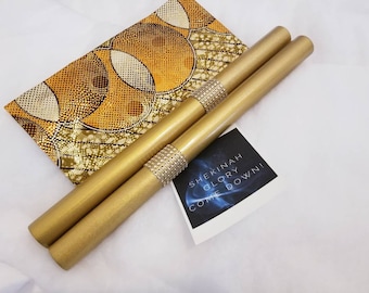 May include: Two cylindrical gold-colored rods with central bands of clear rhinestones, accompanied by a rectangular fabric piece featuring a gold and white circular pattern and a small card with the text SHEKINAH GLORY COME DOWN!