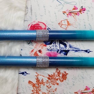 May include: Two cylindrical pens featuring a blue ombre gradient finish that transitions from dark navy to light turquoise. Each pen is accented with a central band of sparkling silver rhinestones, providing a decorative touch to the smooth surface.