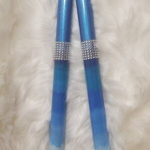 May include: Two cylindrical candles featuring a blue ombre gradient that transitions from a deep metallic blue at the top to a light pale blue at the base. Each candle is accented with a wide band of sparkling silver rhinestones in the center.