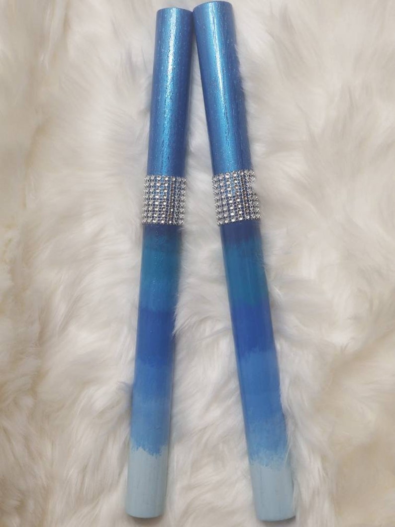 May include: Two cylindrical candles featuring a blue ombre gradient that transitions from a deep metallic blue at the top to a light pale blue at the base. Each candle is accented with a wide band of sparkling silver rhinestones in the center.