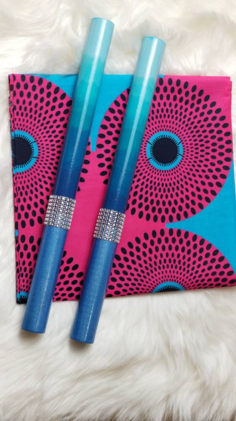 May include: Two cylindrical taper candles featuring a blue ombre gradient from light to dark. Each candle is accented with a band of silver-toned rhinestones around the center. The candles are placed over a square fabric with a pink and blue circular pattern.