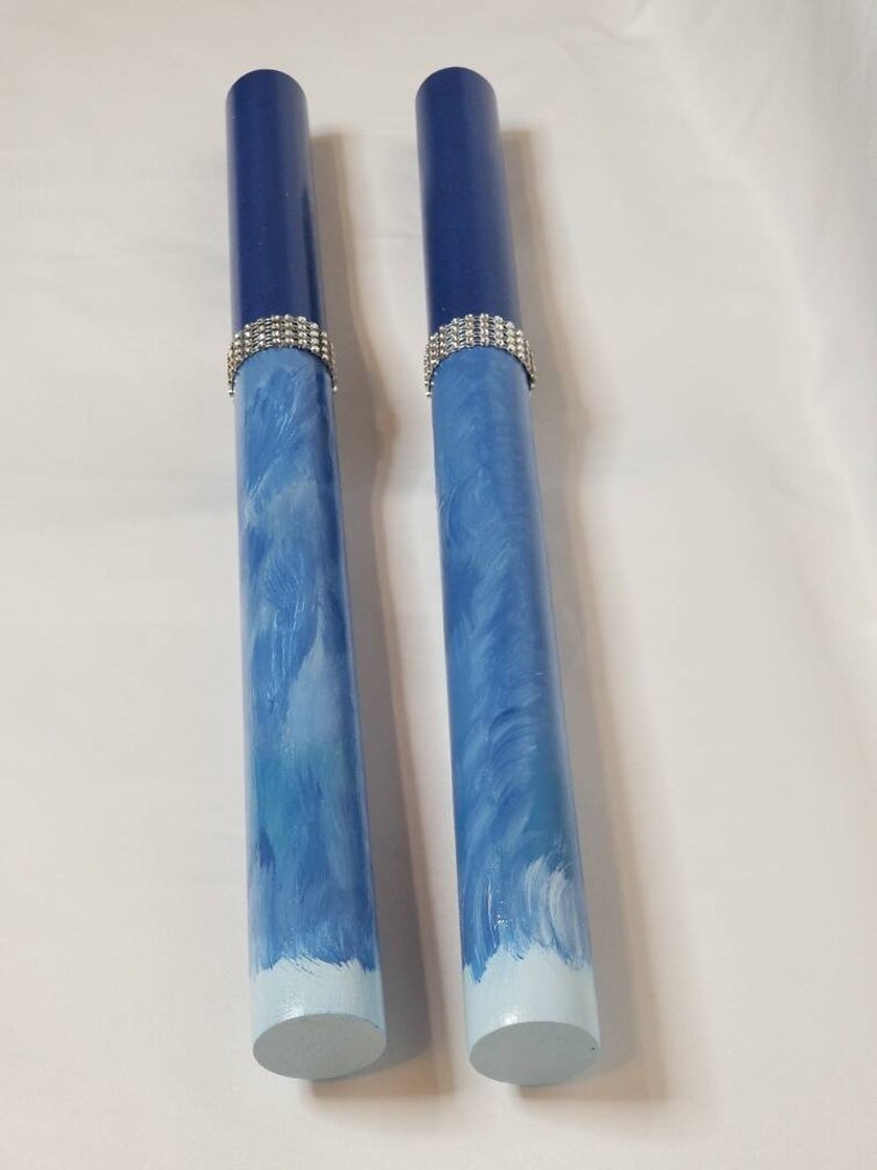 May include: Two cylindrical wooden rods featuring a dark blue top section, a silver-toned rhinestone band, and a light blue base with white brushstroke patterns. The rods have a smooth finish and are positioned vertically side by side.