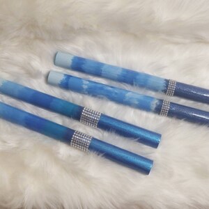 May include: Four cylindrical pens featuring a blue ombre gradient finish that transitions from light to dark blue. Each pen is accented with a band of silver-toned rhinestones positioned near the darker end of the barrel, creating a decorative detail.