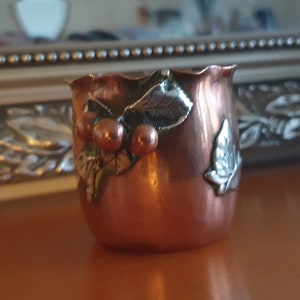 May include: A copper-coloured votive candle holder with a decorative design of cherries and leaves. The leaves are silver-coloured, and the cherries are a darker copper colour. The rim of the holder has a wavy design.