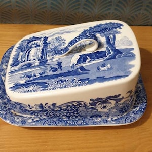 May include: A blue and white ceramic butter dish with a lid featuring a pastoral scene. The lid has a handle and the base has a decorative border.
