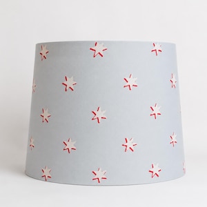 May include: A light blue lampshade with a repeating pattern of red and white stars. The lampshade has a conical shape and is designed to diffuse light. The stars are evenly spaced across the surface of the lampshade.