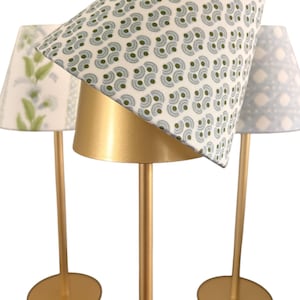 May include: Three decorative table lamps with gold-colored bases and stems. The lampshades feature different patterns: a white and green floral design, a white and green circular pattern, and a light blue geometric pattern. The lamps are designed for indoor use.