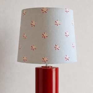 May include: A red ceramic table lamp with a cylindrical base and a light blue lampshade. The lampshade features a repeating pattern of red star shapes. The lamp is a decorative lighting fixture.