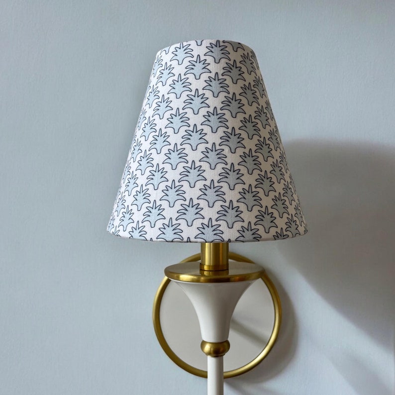 May include: A wall-mounted sconce with a white and gold finish. The lamp shade is cone-shaped and features a repeating pattern of blue leaf-like shapes on a white background. The sconce has a circular gold accent.