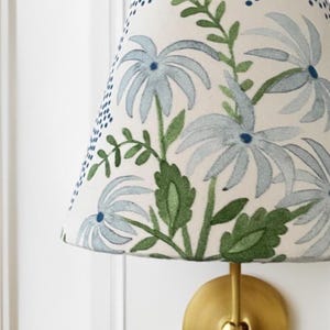 K&ouml;nnte beinhalten: Ein wei&szlig;er Lampenschirm mit einem blauen und gr&uuml;nen Blumenmuster. Der Lampenschirm ist kegelf&ouml;rmig und hat einen goldenen Metallfu&szlig;.