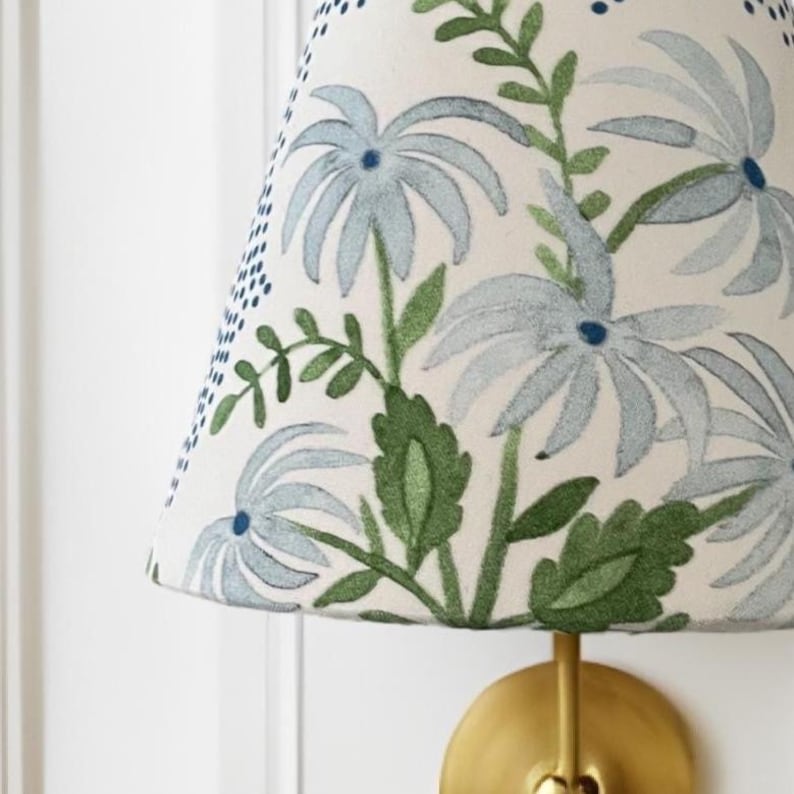 K&ouml;nnte beinhalten: Ein wei&szlig;er Lampenschirm mit einem blauen und gr&uuml;nen Blumenmuster. Der Lampenschirm ist kegelf&ouml;rmig und hat einen goldenen Metallfu&szlig;.