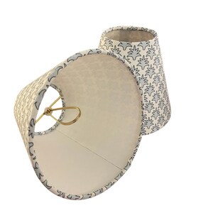 May include: Two small, off-white lampshades with a light blue floral pattern. The shades have a gold-colored metal frame and a white interior. The lampshades are cone-shaped and are designed to be placed on a lamp base.