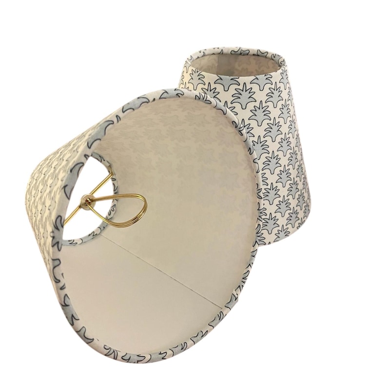 May include: Two small, off-white lampshades with a light blue floral pattern. The shades have a gold-colored metal frame and a white interior. The lampshades are cone-shaped and are designed to be placed on a lamp base.