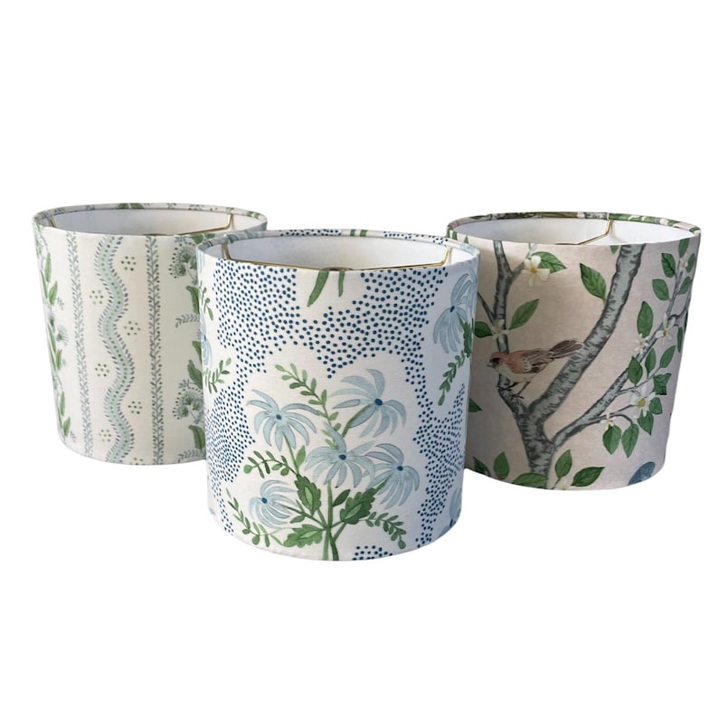K&ouml;nnte beinhalten: Drei zylindrische Lampenschirme mit verschiedenen Blumenmustern. Die Lampenschirme sind wei&szlig; mit blauen und gr&uuml;nen Blumenmustern. Ein Lampenschirm hat ein Muster aus kleinen blauen Punkten, ein anderer hat ein Muster aus blauen Blumen und gr&uuml;nen Bl&auml;ttern und der dritte hat ein Muster aus wei&szlig;en Blumen und gr&uuml;nen Bl&auml;ttern auf einem hellrosa Hintergrund.