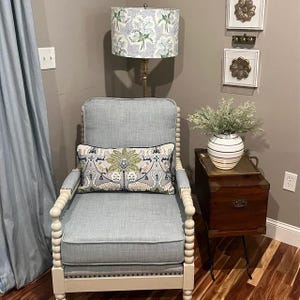 K&ouml;nnte beinhalten: Ein hellblauer Sessel mit einem wei&szlig;en Holzrahmen und Perlendetails. Der Sessel hat ein gemustertes Kissen und steht in einem Raum mit einer Lampe und einem Beistelltisch.