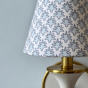 May include: A white and blue patterned lampshade with a brass base. The lampshade has a repeating design of stylized blue leaves on a white background. The lamp is mounted on a light blue wall.