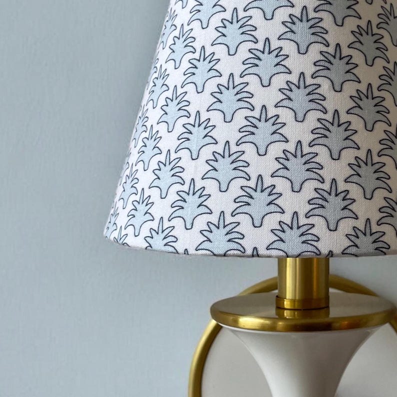 May include: A white and blue patterned lampshade with a brass base. The lampshade has a repeating design of stylized blue leaves on a white background. The lamp is mounted on a light blue wall.