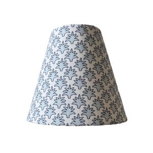 May include: A tapered lampshade with a white background and a repeating pattern of light blue stylized leaf shapes. The lampshade is made of fabric and is designed to fit over a lamp base. The overall shape is conical.