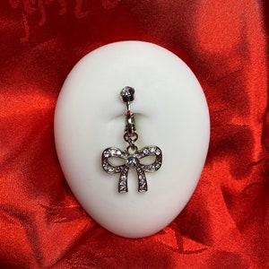 May include: A silver body piercing with a clear crystal and a dangling bow charm decorated with clear crystals.