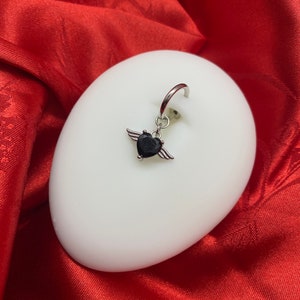 May include: A silver body piercing with a black heart charm and small wings. The charm is attached to a silver ring.