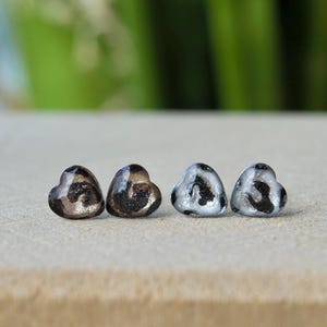 Könnte beinhalten: Vier herzförmige Ohrstecker. Zwei sind dunkel mit glitzernden Akzenten, und zwei sind silberfarben mit schwarzen Mustern. Die Ohrringe sind in einer Reihe auf einer neutralen Oberfläche angeordnet.
