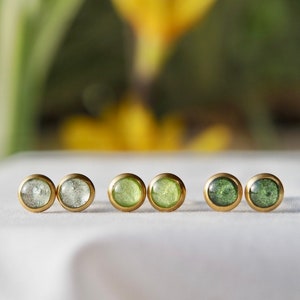 Könnte beinhalten: Sechs goldene Ohrstecker mit grünen und klaren Harzeinsätzen. Die Ohrringe sind in zwei Reihen zu je drei angeordnet.