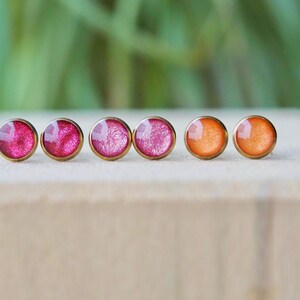 Könnte beinhalten: Sechs Ohrstecker mit goldfarbenen Fassungen. Die Ohrringe haben runde, farbenfrohe, harzgefüllte Mittelstücke. Die Farben umfassen Rosa- und Orangetöne. Die Ohrringe werden auf einer hellen Oberfläche präsentiert.
