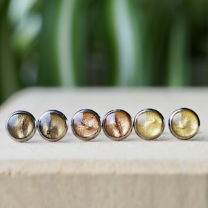 Könnte beinhalten: Sechs kleine, runde, metallische Ohrringe mit goldfarbener Oberfläche. Die Ohrringe haben einen dezenten Schimmer und sind in einer Reihe auf einer Holzoberfläche angeordnet.