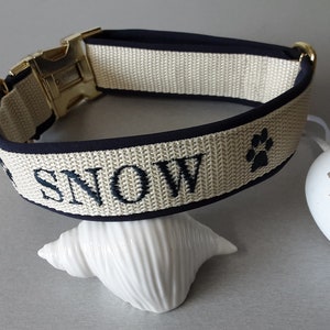 May include: A beige dog collar with a black trim and gold buckle. The collar is embroidered with the name "SNOW" and a paw print.