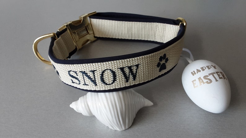 May include: A beige dog collar with a black trim and gold buckle. The collar is embroidered with the name "SNOW" and a paw print.