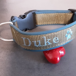 May include: A gold and blue dog collar with the name "Duke" embroidered on it. The collar has a silver D-ring and a black plastic buckle.