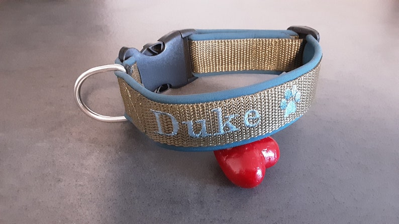May include: A gold and blue dog collar with the name "Duke" embroidered on it. The collar has a silver D-ring and a black plastic buckle.