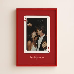 Puede incluir: Un cuadro con marco rojo que presenta un diseño de carta de juego as de corazones. El centro de la carta muestra una fotografía de una pareja. Debajo de la carta, el texto "how lucky are we" está escrito en una fuente cursiva blanca.