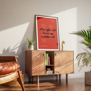 Puede incluir: Una impresi&oacute;n enmarcada con fondo rojo y texto negro. El texto dice "And after all you're my wonderwall". La impresi&oacute;n est&aacute; colgada en una pared sobre un armario de madera con una planta en una maceta en el lado izquierdo del armario.