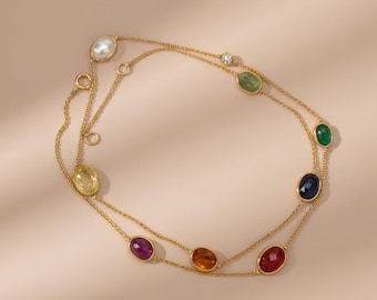 May include: A delicate gold necklace featuring a double strand design. The necklace is adorned with a variety of colorful gemstones, including a pearl, emerald, sapphire, and ruby. The gemstones are oval-shaped and set in gold bezels.