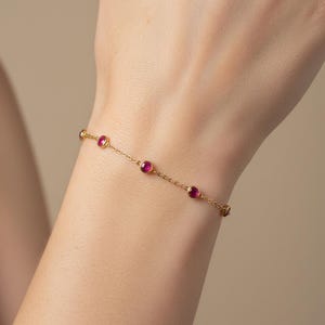 May include: A delicate gold bracelet featuring small, round, ruby-colored gemstones. The bracelet is worn on a wrist, showcasing the fine chain and the vibrant red of the gemstones. The bracelet is a piece of jewelry.