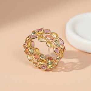 以下が含まれることがあります： 金色のリング。 涙の形をしたカラフルな宝石が繰り返されるパターン。 宝石は黄色、ピンク、オレンジ、緑の色合いです。 リングは、柔らかいピーチ色の背景に配置されています。