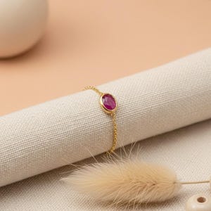 May include: A fine gold bracelet with a vivid, oval-cut ruby gemstone. The ruby is set in a gold bezel and attached to a delicate gold chain. The bracelet is presented on a neutral fabric, with a soft, blurred background.