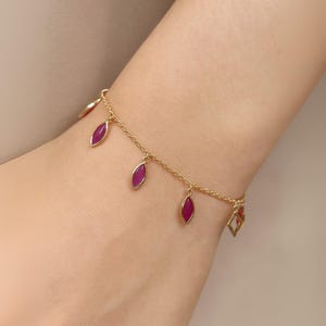 May include: A gold-colored bracelet with dangling magenta-colored leaf-shaped charms. The bracelet is delicate and appears to be made of thin chain links. The charms are evenly spaced along the chain, adding a touch of elegance to the jewelry.
