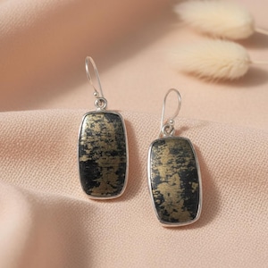 Può includere: Un paio di orecchini pendenti argentati. Ogni orecchino presenta una pietra nera rettangolare con accenti dorati strutturati. Gli orecchini pendono da un semplice gancio e sono esposti su una superficie rosa.