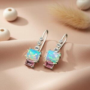 May include: A pair of silver drop earrings with iridescent, rectangular opal-like stones and small, round clear stones. Each earring also features a rectangular pink stone at the bottom. The earrings are displayed on a pink fabric.