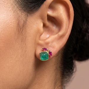Pode incluir: Um close-up de um brinco dourado com uma pedra quadrada de cor esmeralda e duas pedras menores de cor rubi. O brinco é colocado em uma moldura dourada e usado em uma orelha.