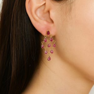 May include: Gold chandelier earrings with teardrop-shaped, ruby-colored gemstones. The earrings feature a delicate, layered design with multiple gemstones suspended from a central stud. The gemstones are a vibrant red hue.